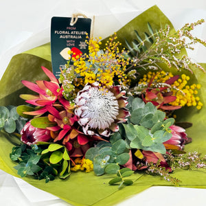 A beautifully arranged Australian Native Flower Bouquet featuring a large King or Queen Protea from the Adelaide Hills as the centerpiece. Surrounding the protea are seasonal native blooms in soft pastel or vibrant tones, artistically wrapped in elegant paper and tied with a delicate ribbon. The bouquet showcases a natural, organic style with a mix of textures and colors, perfect for any special occasion.