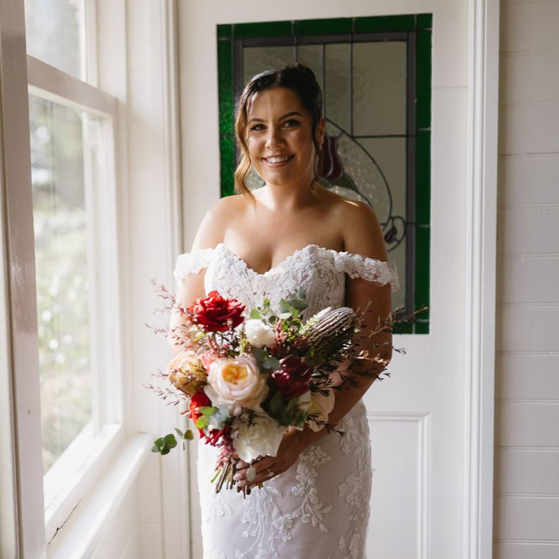 Autumn Grace Bridal Bouquet by Floral Atelier Australia – hand-tied bouquet with red Ecuador roses, cream Grandiflora roses, and seasonal native flowers like banksia and protea in rich autumn tones.