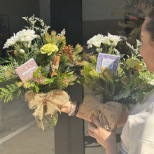 Load image into Gallery viewer, Person holding two Christmas bouquets of flowers with decorative ribbons in front of a door.
