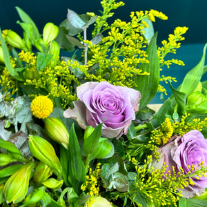 A close-up image of a stunning pastel rose arrangement featuring pink and purple roses with delicate lavender accents. The vibrant roses and lush green foliage create a soft, elegant look, highlighting the fine details of the fresh blooms in this beautiful floral display.