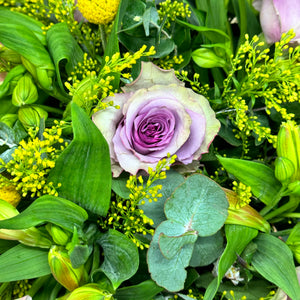 A close-up image of a stunning pastel rose arrangement featuring pink and purple roses with delicate lavender accents. The vibrant roses and lush green foliage create a soft, elegant look, highlighting the fine details of the fresh blooms in this beautiful floral display.