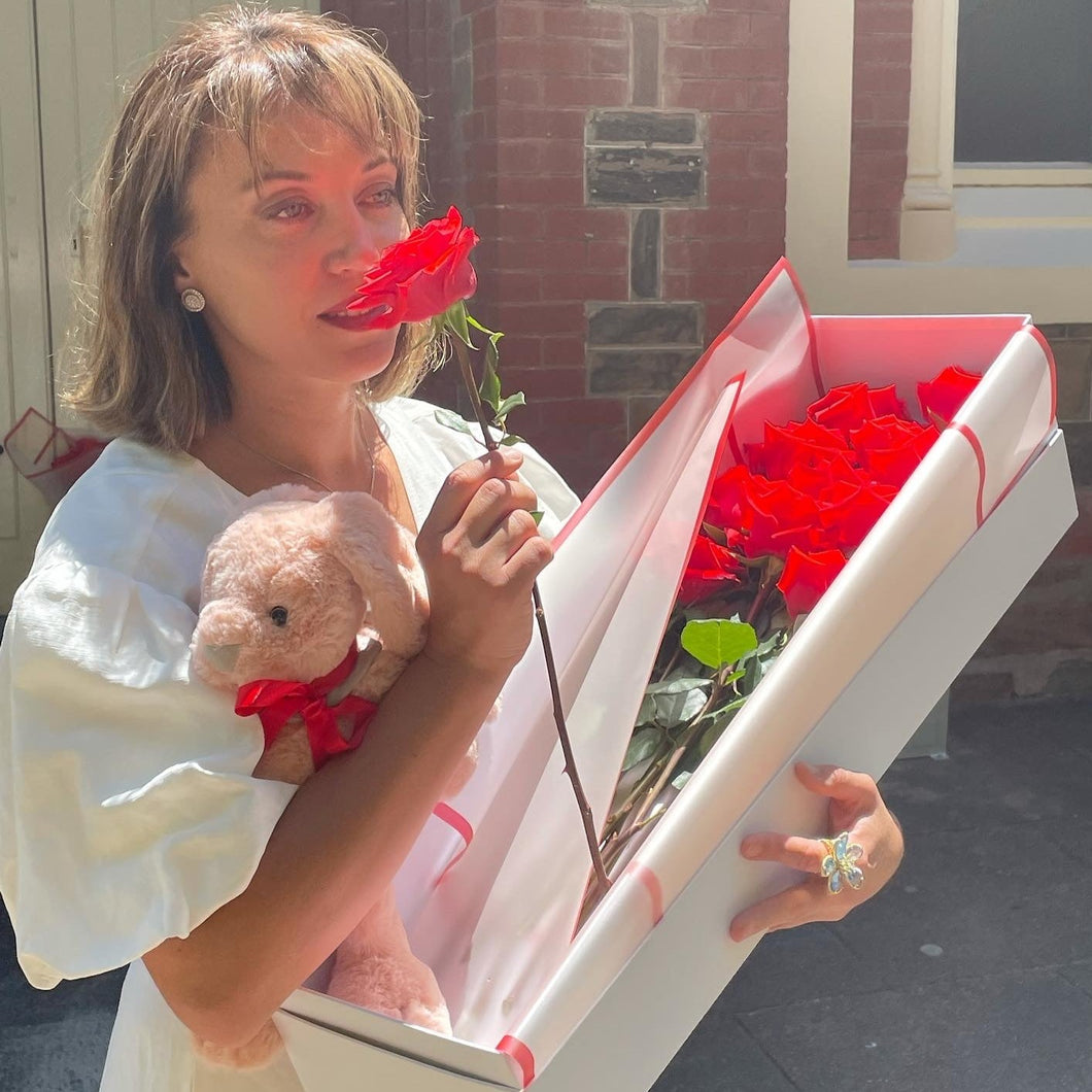 A heartfelt moment as a woman beholds a Dozen Ferrari Red Roses with a charming plush bunny, beautifully boxed by Floral Atelier Australia, ready to enchant with same day flower delivery in Adelaide