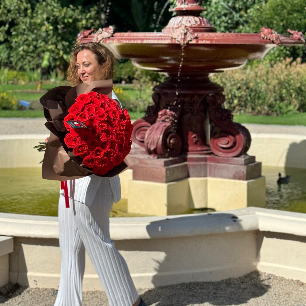 In the tranquil Adelaide Botanic Garden, a joyous woman cherishes Floral Atelier Australia's Elegant 50 Ferrari Red Roses Bouquet, a luxurious and captivating symbol of affection, available for same day flower delivery from the finest florist in Adelaide