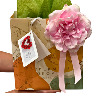 A stylish gift bag from Floral Atelier Australia, presented in a Peppermint Grove Australia bag decorated with a large pink silk flower and pink ribbon. The bag features soft beige, orange, and green floral patterns and is filled with green tissue paper. A Valentine’s Day card with a heart made of red roses is attached to the bag.