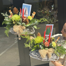 Load image into Gallery viewer, Decorative floral Christmas arrangement with candles and pinecones on a stand, held by a person.
