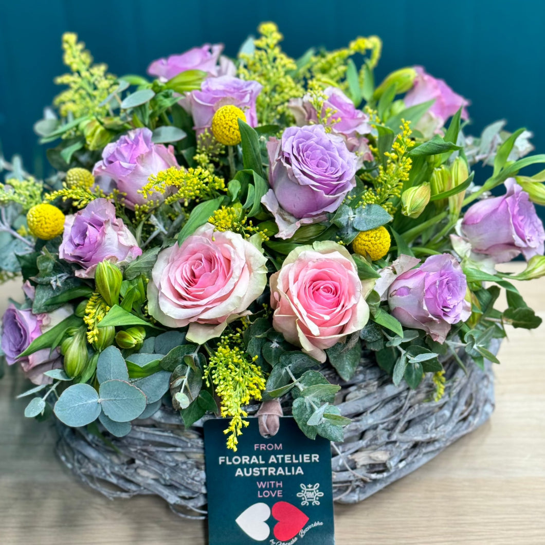 A lavish pastel rose arrangement in a glass vase with 2 pink roses, 10 purple roses, 5 alstroemerias, 4 yellow billy buttons, and rich foliage of golden rod and blue gum. The deluxe size showcases a fuller, more vibrant display, ideal for making a bold and elegant statement.