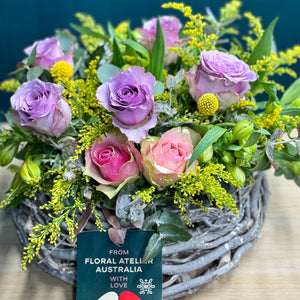 A beautiful pastel rose arrangement in a glass vase featuring 2 pink roses, 5 purple roses, 5 alstroemerias, 2 yellow billy buttons, and lush accents of golden rod and blue gum. The standard size offers a harmonious blend of soft colors and textures, perfect for adding a touch of elegance to any space.