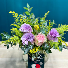 Load image into Gallery viewer, A delicate pastel arrangement in a glass vase featuring 1 pink rose, 3 purple roses, a yellow billy button, and accents of golden rod and blue gum. The compact size showcases subtle elegance and gentle beauty, perfect for smaller spaces or thoughtful gestures.
