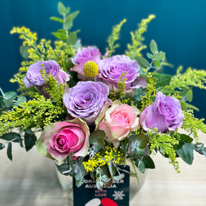 A fuller and vibrant pastel rose posy displayed in a glass vase, containing 2 pink roses, 5 purple roses, a yellow billy button, and lush accents of golden rod and blue gum. This arrangement offers a more generous display of soft, soothing colors, ideal for making a warm and welcoming statement.