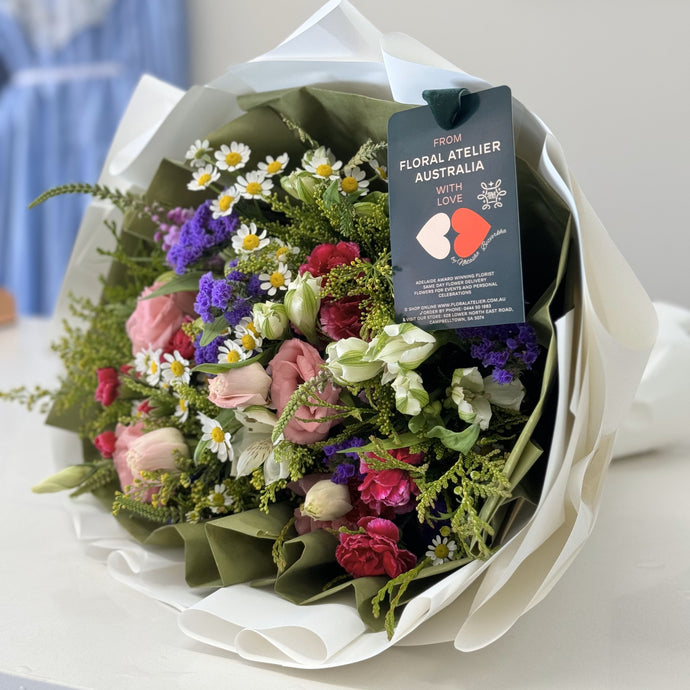 A vibrant floral arrangement featuring a mix of seasonal flowers in blue, purple, and violet hues. The bouquet is composed of delicate blooms and lush greenery, creating a bold and colorful display that conveys elegance and joy. Arranged in a compact but full design, this bouquet exudes sophistication and charm.