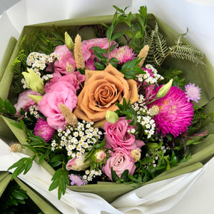 A beautiful floral arrangement featuring a unique toffee Ecuador rose at the center, surrounded by seasonal pink lisianthus, roses, delicate white daisies, and textured blooms, complemented by aromatic pine foliage. The bouquet is arranged in a compact but full design, creating a stunning and memorable display.