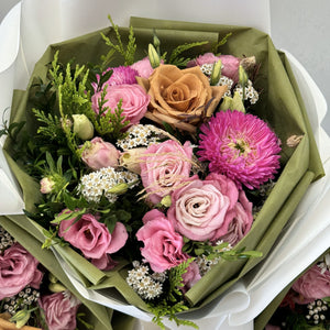 A beautiful floral arrangement featuring a unique toffee Ecuador rose at the center, surrounded by seasonal pink lisianthus, roses, delicate white daisies, and textured blooms, complemented by aromatic pine foliage. The bouquet is arranged in a compact but full design, creating a stunning and memorable display.