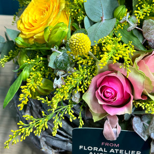 A close-up view of the Sunshine Rose Arrangement, highlighting the vibrant pink and yellow roses, delicate alstroemerias, and cheerful yellow billy buttons, accented with golden rod and lush blue gum foliage. The detailed shot captures the fresh, radiant colors and intricate textures of the flowers, showcasing the bouquet's lively and elegant composition.