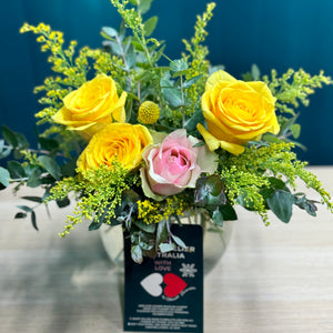 A delightful mini arrangement of yellow and pink roses, featuring a single pink rose surrounded by three bright yellow roses, accented with a yellow billy button and lush greenery, all elegantly arranged in a small glass vase.