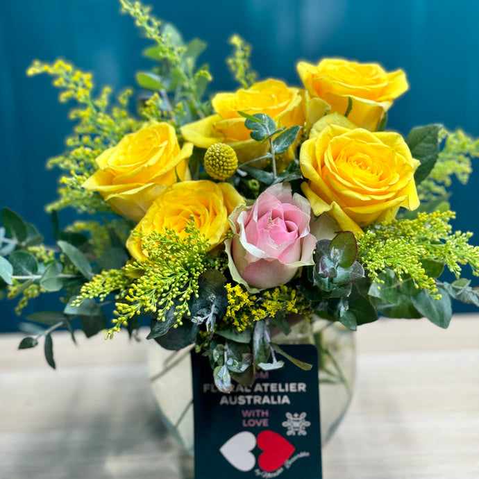 A cheerful standard arrangement of yellow and pink roses, featuring two pink roses surrounded by five vibrant yellow roses, accented with a yellow billy button and lush greenery, all elegantly arranged in a medium-sized glass vase.