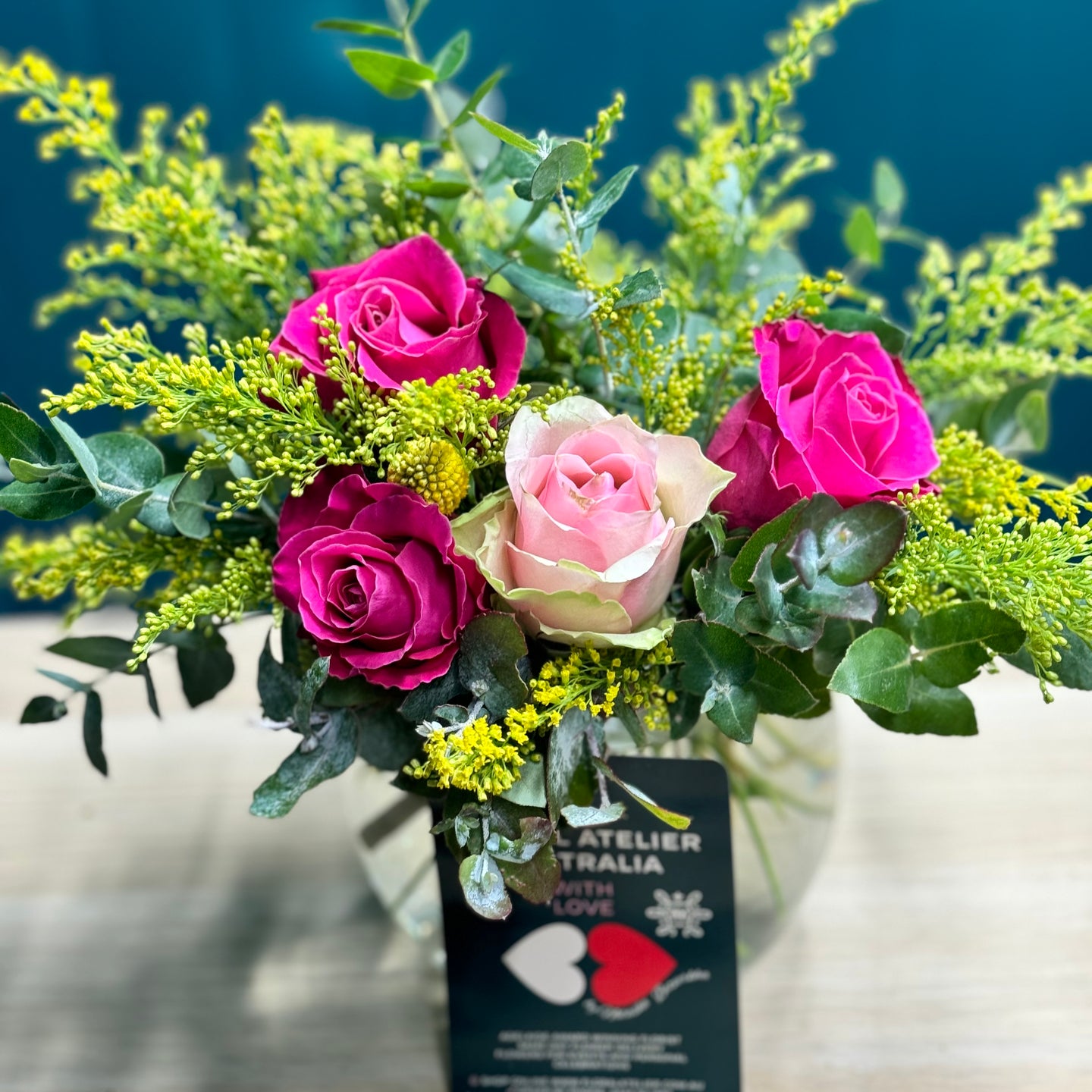 A vibrant rose posy in a glass vase featuring 1 pink rose, 3 hot pink roses, 1 yellow billy button, and lush accents of golden rod and blue gum. The mini size offers a compact yet lively display, perfect for adding a pop of color and cheer to any space.