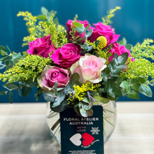 Load image into Gallery viewer, A beautiful vibrant rose posy in a glass vase with 2 pink roses, 5 hot pink roses, 1 yellow billy button, and rich greenery of golden rod and blue gum. The standard size showcases a fuller arrangement with bright, joyful colors, ideal for making a cheerful statement.

