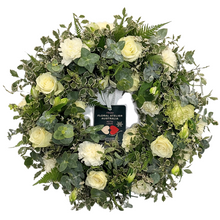 Load image into Gallery viewer, A medium-sized white floral wreath made with roses and carnations, surrounded by green foliage.
