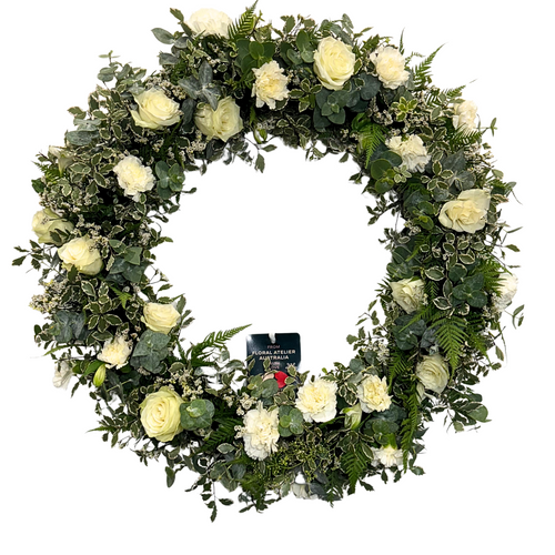 A medium-sized white floral wreath made with roses and carnations, surrounded by green foliage.