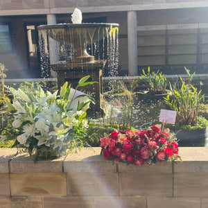 Red Rose Wreath and Pure Life Arrangement Deluxe placed together at Pasadena Cemetery, highlighting their combined beauty amidst a serene outdoor setting with a waterfall backdrop.